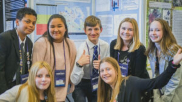 High school students at the Canada-Wide Science Fair standing in front of a project board, smiling at the camera and waving