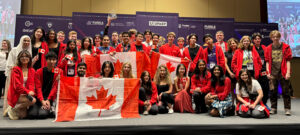 A large group of young people wearing red and white, with many holding Canadian flags, posing for a photo at MILSET ESI 2024. They are standing and sitting in several rows, smiling and looking at the camera. In the background, there are banners with logos.