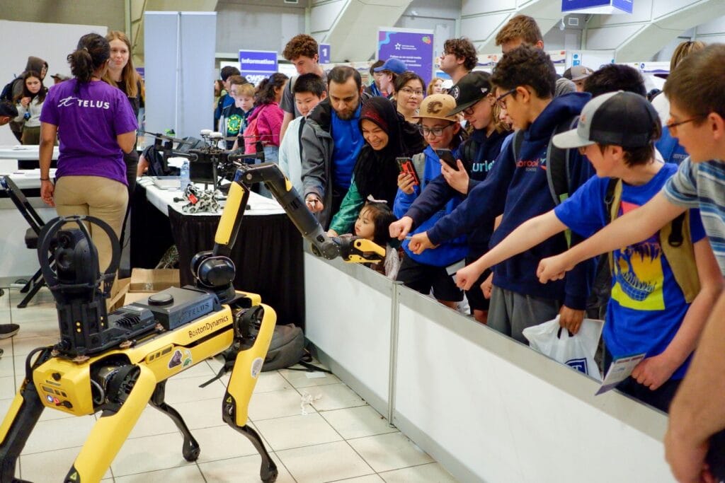 The Telus booth at the Canada-Wide Science Fair STEM Expo. In the foreground, a four-legged yellow and black robot, labeled Boston Dynamics, captivates onlookers. People of varying ages, including children in blue shirts and adults, watch intently, some taking photos.