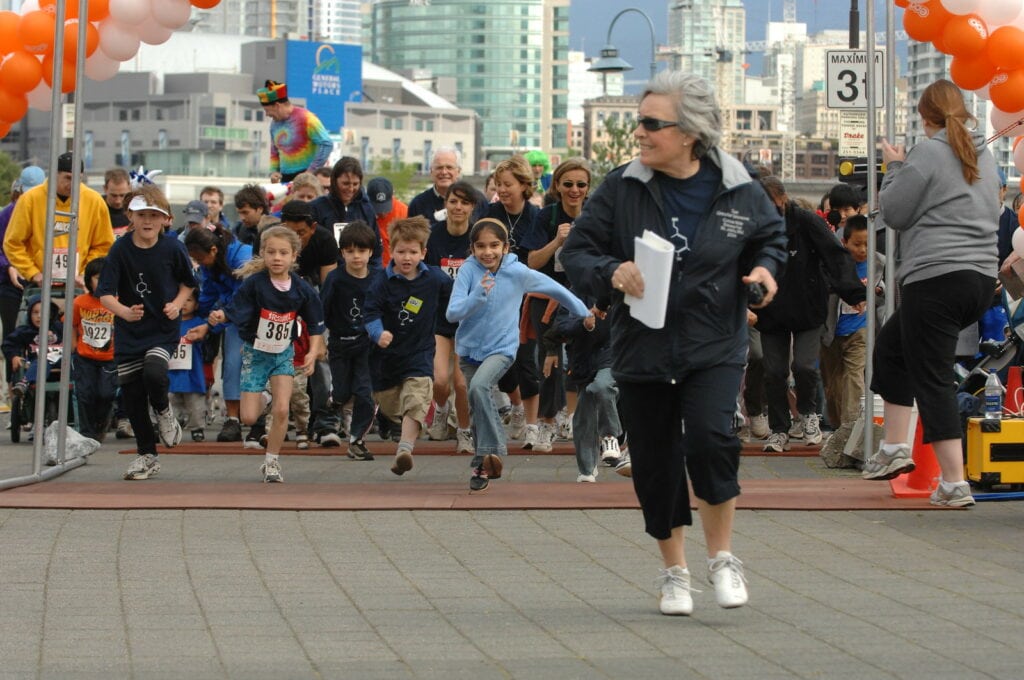 Children and youth participating in a science fair event, running towards the finish line with a supervising adult, in an urban outdoor setting with city buildings in the background.