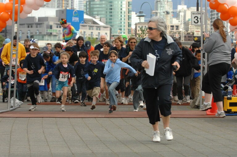 Children and youth participating in a science fair event, running towards the finish line with a supervising adult, in an urban outdoor setting with city buildings in the background.