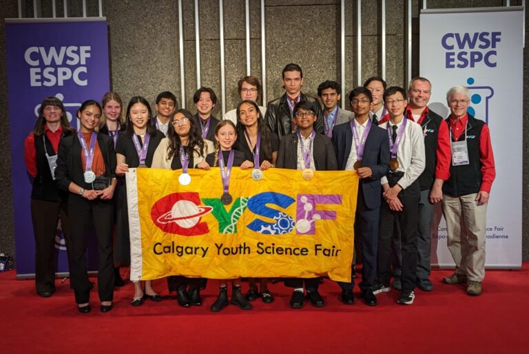 Group of diverse youth participants at the Calgary Youth Science Fair holding a colorful banner, with organizers and sponsors in the background, celebrating science and innovation.