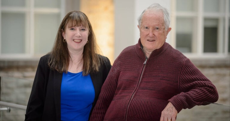 Two smiling individuals, a young woman and an older man, standing outdoors in front of a building, representing Youth Science Canada and promoting youth engagement in science activities.