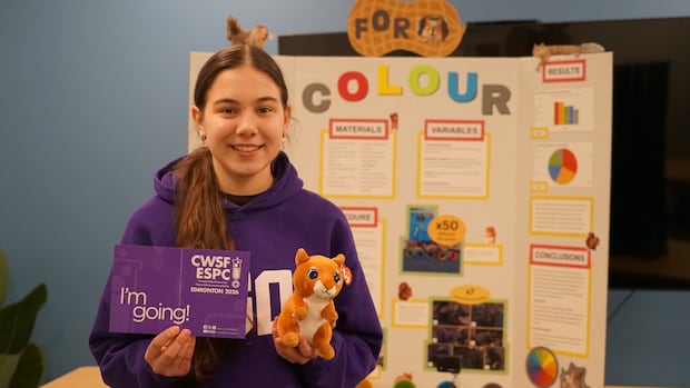 A young girl presenting her science project at Youth Science Canada event, holding a purple sign and a plush toy, with a colorful research poster in the background.