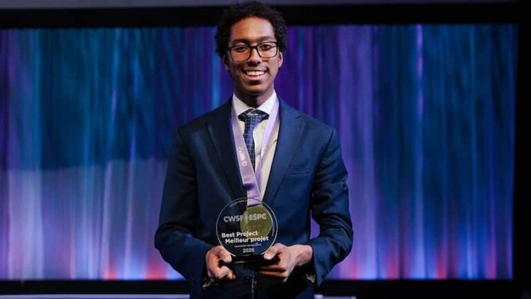 Young man in formal suit holding an award at Youth Science Canada event, smiling on stage with colorful curtain background.