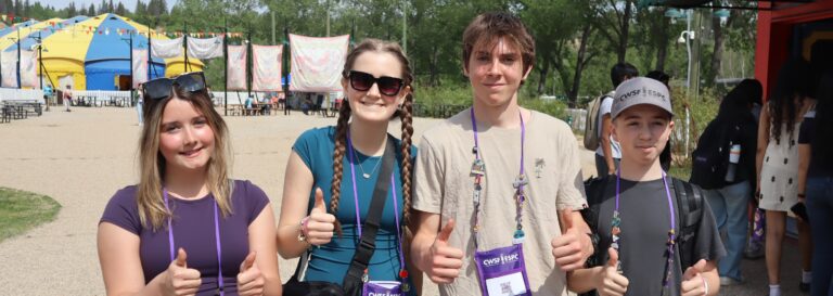 Group of four young students at Youth Science Canada event, smiling and giving thumbs up, with tents and trees in the background, promoting STEM education and youth engagement.