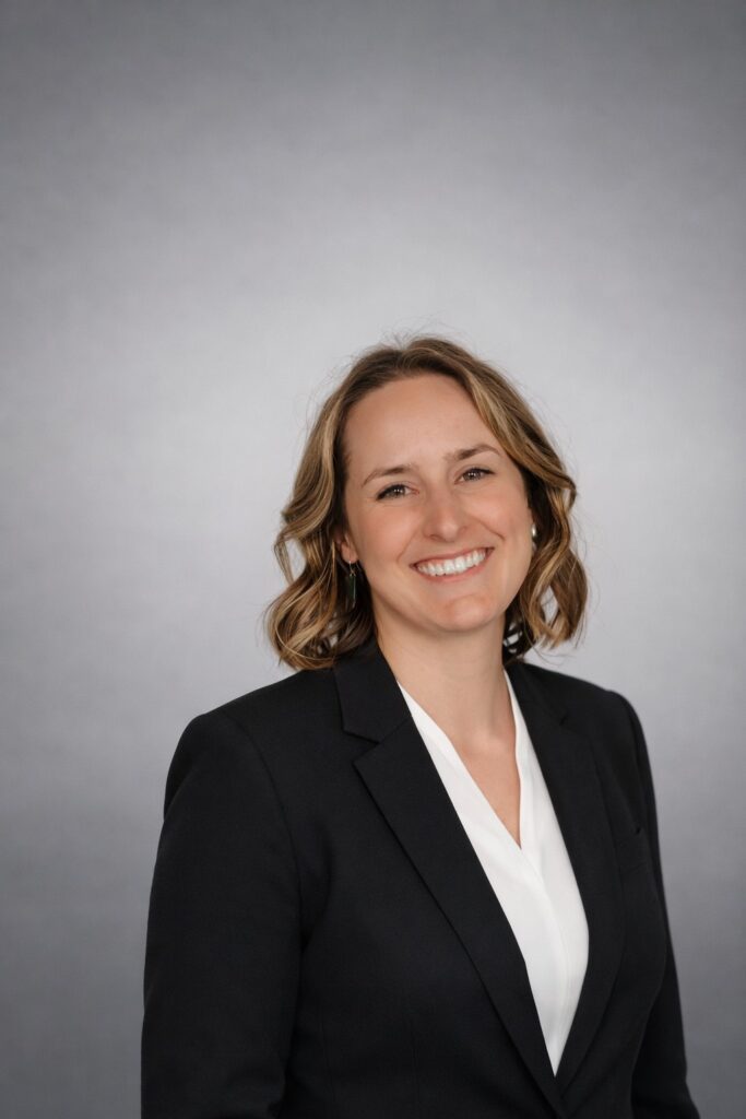 Young woman with shoulder-length wavy hair smiling in a professional black blazer and white shirt against a gray background.