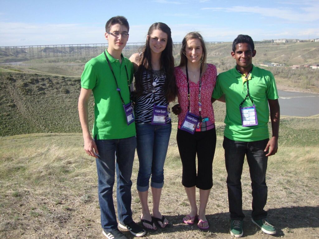 Group of four young students at a science fair outdoors, wearing badges, with a scenic landscape and river in the background.