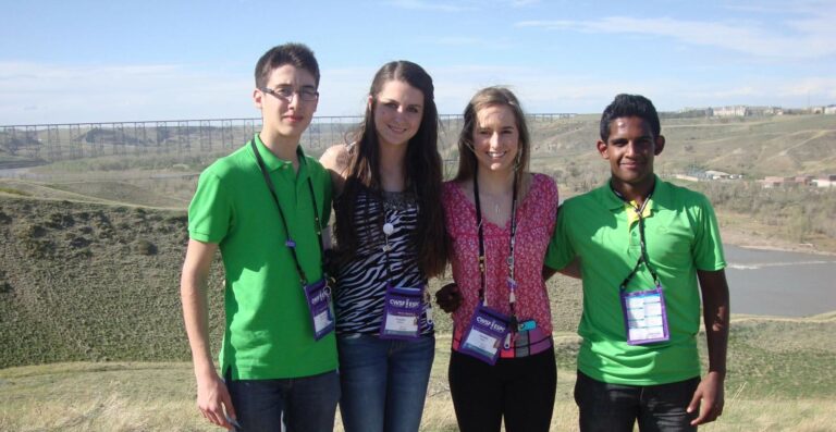 Group of four young students at a science fair outdoors, wearing badges, with a scenic landscape and river in the background.